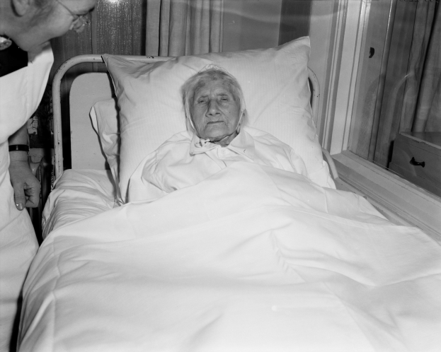 Eine ältere Frau liegt in einem Krankenhausbett mit einer weißen Decke und einem Kissen, während eine Krankenschwester neben ihr steht, mit einem Fenster mit Vorhängen im Hintergrund.