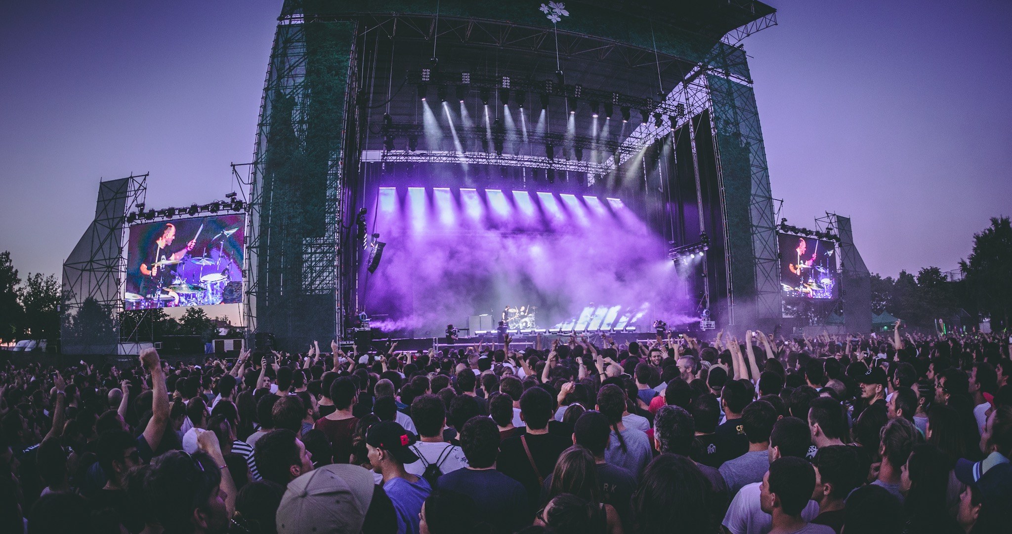 Eine große Menschenmenge auf einem Musikfestival mit einer Bühne, auf der Musiker spielen, beleuchtet von Scheinwerfern und umgeben von Bildschirmen, vor einer Kulisse aus einem Gebäude, Bäumen und einem klaren blauen Himmel.