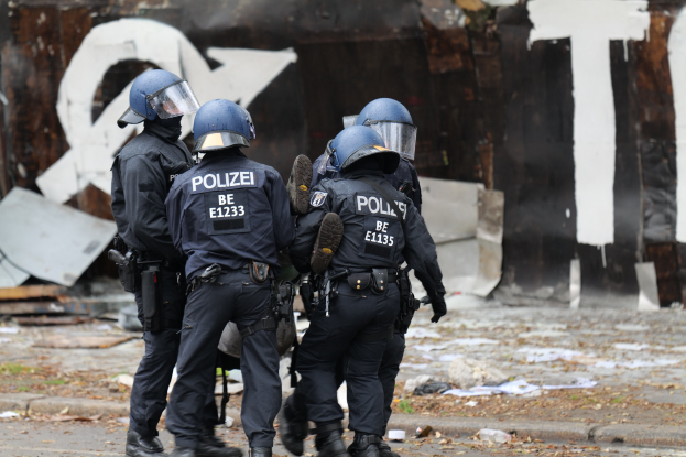 Polizeibeamte in schwarzen Uniformen und blauen Helmen stehen vor einem graffiti-bedeckten Gebäude, umgeben von verstreuten Papieren und Trümmern, mit einer textbedeckten Wand im Hintergrund.