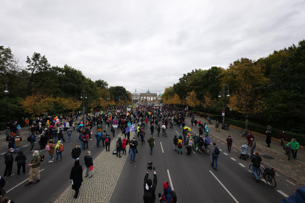 Große Gruppe von Menschen marschiert eine baumbestandene Straße in Berlin entlang, einige halten Kameras, mit einem Gebäude und einem klaren Himmel im Hintergrund.