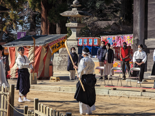 Eine Gruppe von Menschen in traditioneller Kleidung versammelt sich im Freien in Kyoto, einige tragen Masken und halten Holzstöcke, mit Stühlen, Bannern und einem Zelt gegen einen klaren blauen Himmel.
