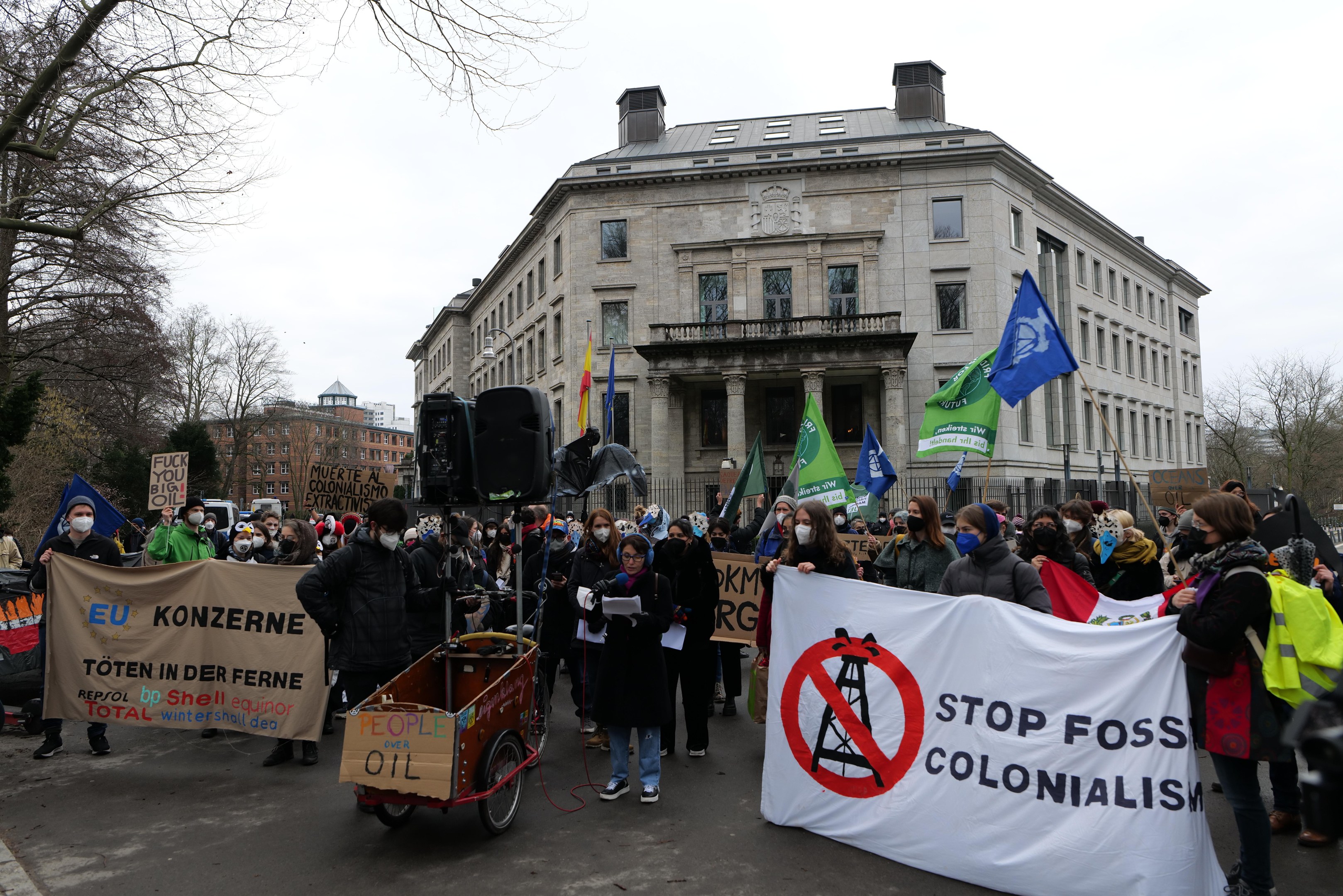Große Gruppe von Menschen marschiert auf einer Straße bei einer Protestaktion gegen fossile Brennstoffe, trägt Schilder und Fahnen, mit einem Fahrzeug im Vordergrund und Gebäuden, Bäumen und einem klaren blauen Himmel im Hintergrund.