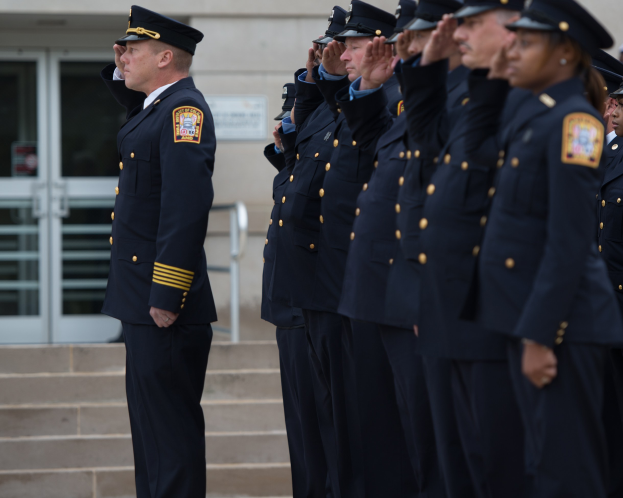 Gruppe von Polizisten in Uniform und Mützen, die in Formation vor einem Gebäude mit Glastüren und Treppe salutieren.