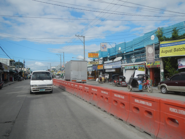 Eine belebte Stadtstraße mit Fahrzeugen, einer Verkehrsinsel, Gebäuden mit Schildern, Strommasten und -leitungen, Bäumen und einem bewölkten Himmel.