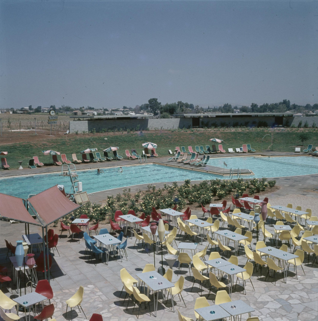 Ein Freibad mit Tischen, Stühlen und Sonnenschirmen, umgeben von Pflanzen, einem Zaun, einer Wand, Bäumen und Gebäuden unter einem bewölkten Himmel.