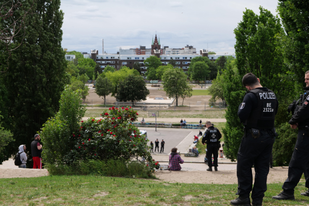 Zwei Polizeibeamte vor einer Gruppe von Menschen in einem Park mit grünem Gras, Bäumen, bunten Blumen, Gebäuden, Pfählen und einem klaren blauen Himmel.