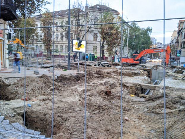 Baustelle mit großer Aushebung umgeben von einem Metallzaun, benachbarte Straße mit Fahrzeugen, Fußgänger auf einem Gehweg, Verkehrsampeln, Schilder, Bäume, Gebäude mit Fenstern und einen klaren blauen Himmel.