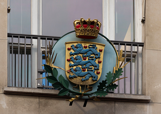 Wappen der britischen Königsfamilie an der Außenseite eines Gebäudes dargestellt, mit einer Krone und einem Blatt-Logo, sowie Fenstern und Metallgittern im Hintergrund.