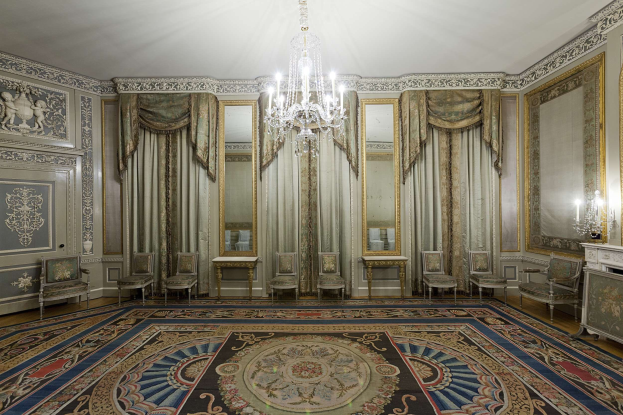 Ein luxuriöses Zimmer mit einem großen Teppich, Stühlen, Tischen, Spiegeln, Vorhängen, einem Kronleuchter und einem Kamin auf der rechten Seite, mit kunstvoll verzierten Wänden.