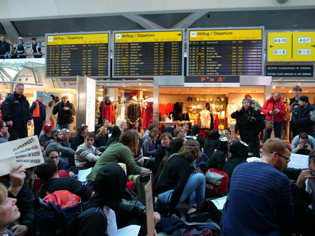 Eine große Gruppe von Menschen in einem Flughafen, einige sitzen mit Taschen und Papieren, andere stehen, mit Texttafeln, Schaufensterpuppen in Kleidern und Deckenbeleuchtung, was auf eine Protestaktion hindeutet.