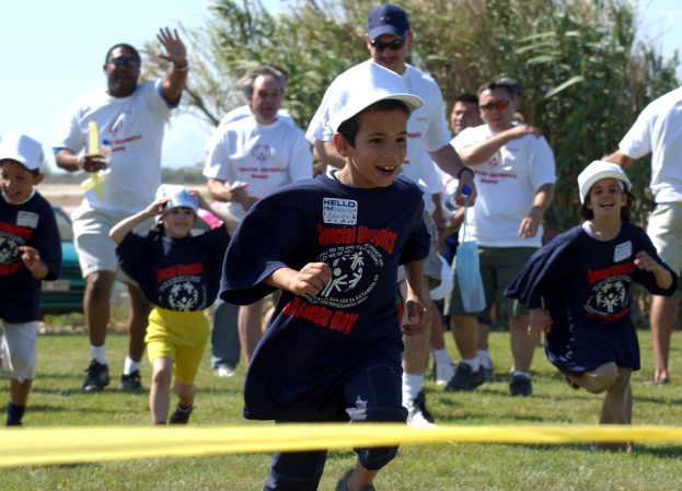 Kinder laufen in einem Rennen auf einem grünen Feld mit einem gelben Band im Vordergrund, einige tragen Kappen und Schutzbrillen, andere halten Gegenstände, mit Bäumen und einem klaren blauen Himmel im Hintergrund.