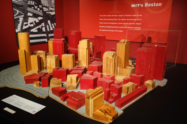 Ein hölzernes Blockmodell einer Stadt auf einem Tisch in einem Museum, begleitet von Papieren, einer Glasbox und einem gerahmten Foto an der Wand.