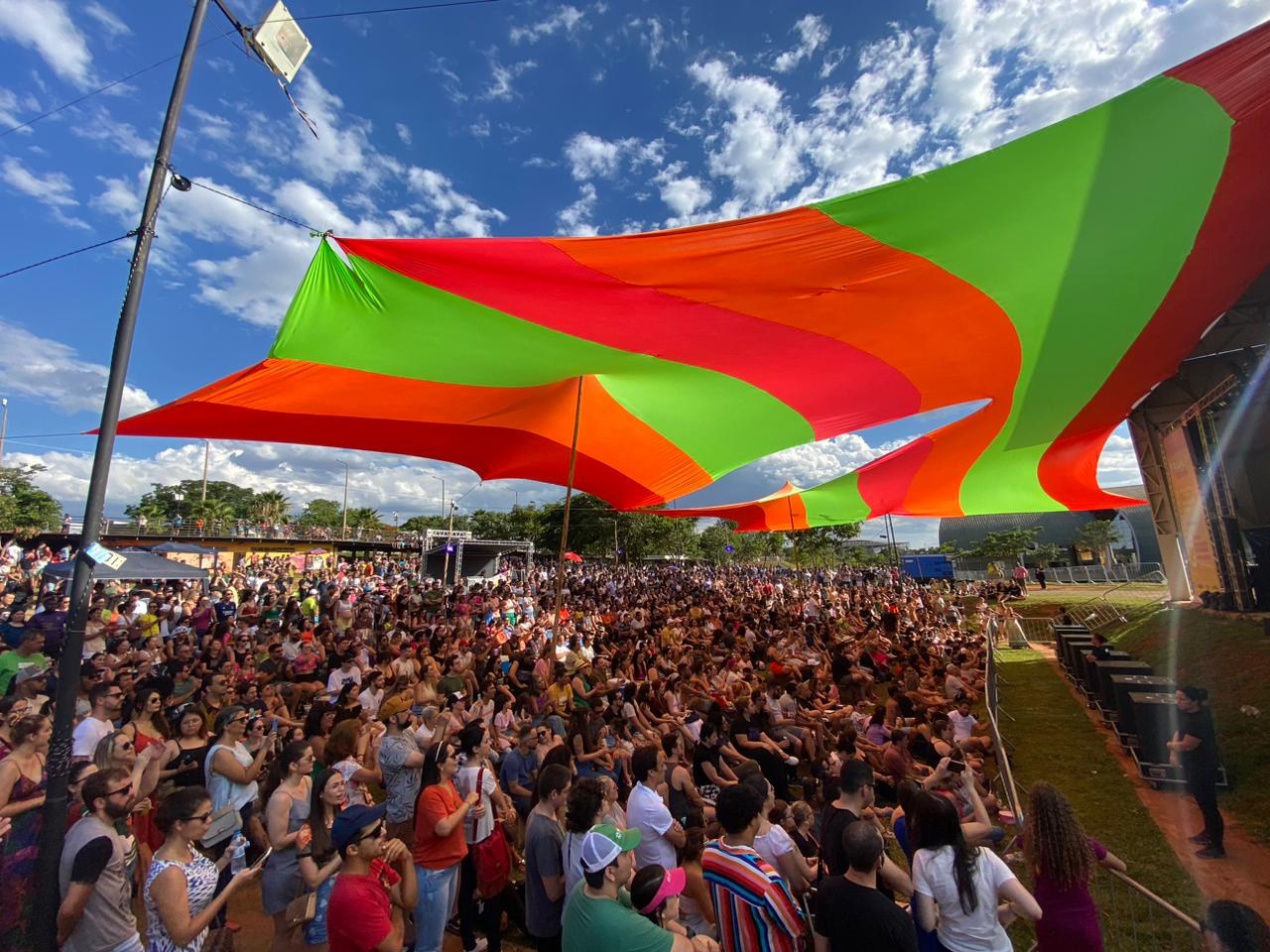 Eine große Menschenmenge steht vor einer Regenbogenflagge, mit einem Gebäude auf der rechten Seite, Lautsprechern auf einer Bühne und Bäumen und Wolken am Himmel während einer Festival-Jubiläumsfeier.