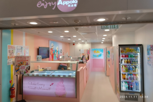 Innenansicht eines Eisdielenladens mit einem Kühlschrank auf der rechten Seite, der mit Flaschen gefüllt ist, einem Feuerlöscher und einer Tafel mit Text auf der linken Seite, einer Person, die an einem Tisch in der Mitte sitzt, und einem Schild mit der Aufschrift 'Genießen Sie das Apollo-Eiscafé' oben.