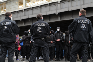 Eine Gruppe von Polizisten in Uniform steht vor einer Menge von Menschen in schwarzen Uniformen und Masken, mit einer Stadtbrücke und einem Gebäude im Hintergrund, während einer Demonstration.
