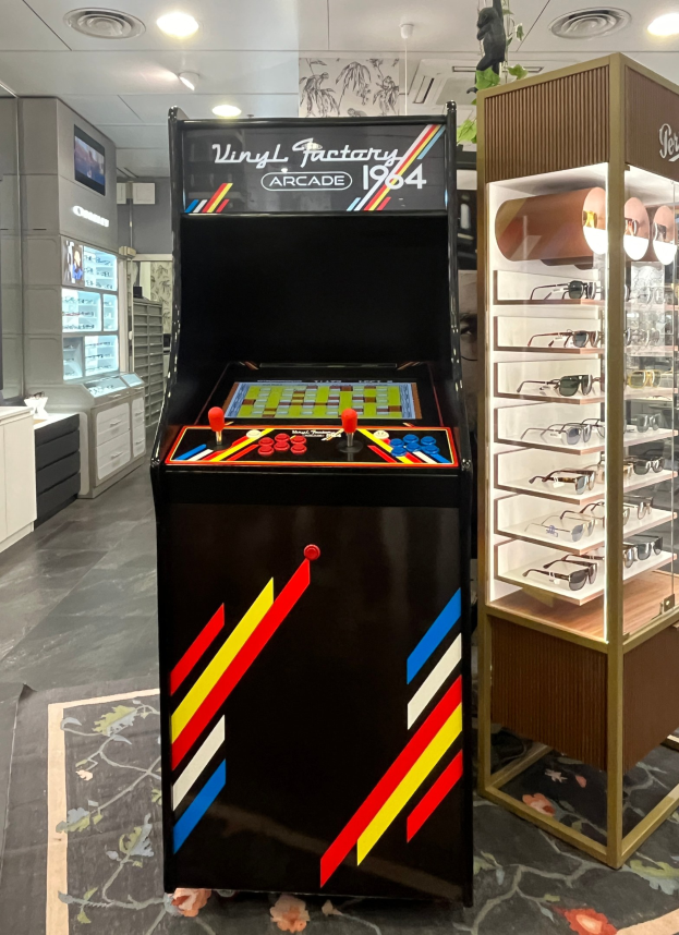 Ein Geschäft mit einem zentralen Retro-Arcade-Automaten, einem Glasvitrinenschrank auf der rechten Seite und Retro-Beleuchtung mit Schränken, Fernseher und anderen Gegenständen im Hintergrund.