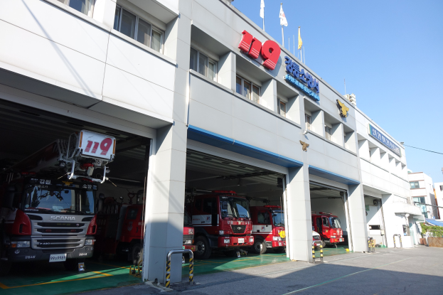 Feuerwache mit mehreren Feuerwehrautos vor dem Gebäude geparkt, mit Fenstern, Säulen und Namensschildern, sowie Flaggen im Vordergrund und anderen Gebäuden unter einem klaren blauen Himmel.