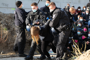 Eine Gruppe maskierter Personen in Mützen, einige mit Waffen, umgibt eine Frau, die von der Polizei eskortiert wird, mit Pflanzen, Steinen und einer Wand mit Protesttext im Hintergrund.