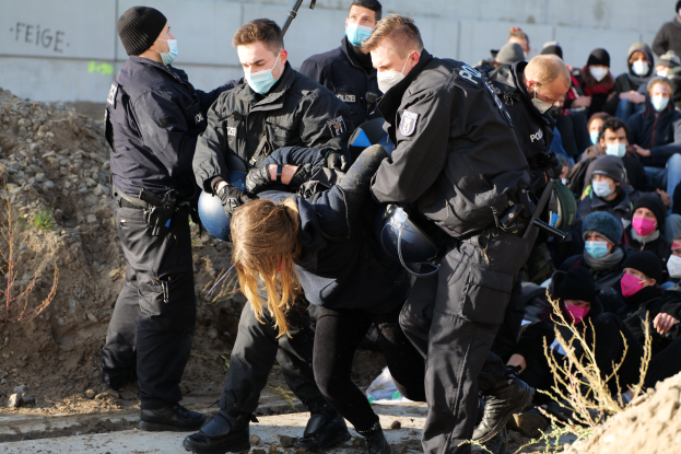 Eine Gruppe maskierter Personen in Mützen, einige mit Waffen, umgibt eine Frau, die von der Polizei eskortiert wird, mit Pflanzen, Steinen und einer Wand mit Protesttext im Hintergrund.
