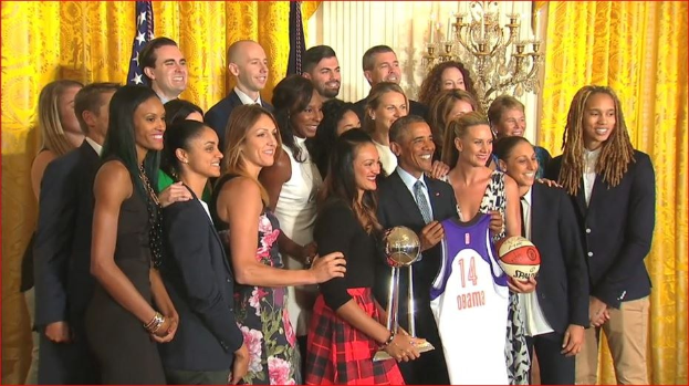 Präsident Obama und First Lady Michelle Obama posieren mit dem Frauen-Basketball-Team im Oval Office des Weißen Hauses, halten einen Basketball, eine Trophäe und lächeln mit einer Flagge, Vorhängen und einem Kerzenleuchter im Hintergrund.