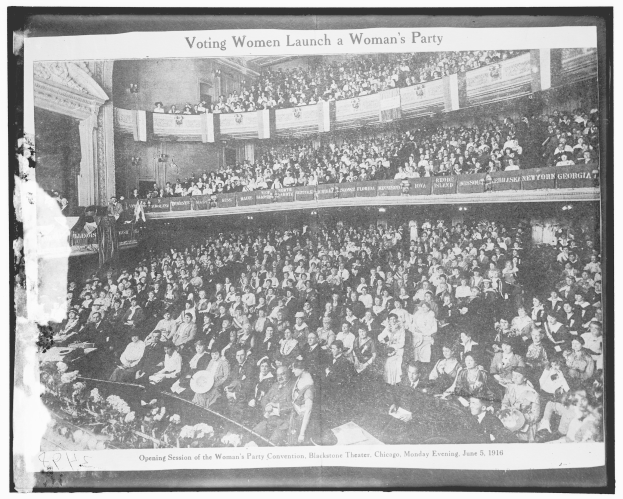 Ein Schwarz-Weiß-Foto einer großen Menschenmenge in einem Gebäude, mit einigen sitzenden und stehenden Personen, Schildern mit Text und einer Wand im Hintergrund. Unten steht der Text "Wahlrecht für Frauen gründet eine Frauenpartei."