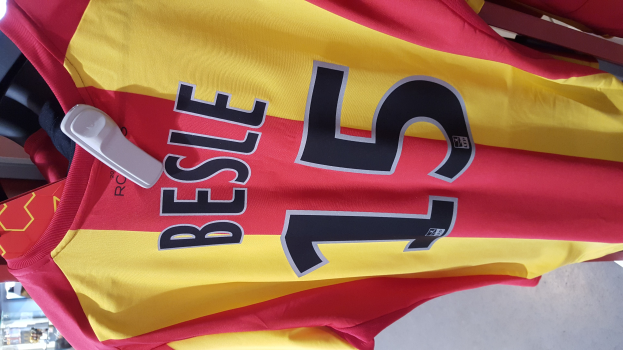 Rotes und gelbes Fußballtrikot mit Text und Zahlen, das an einer Ladengestell hängt, mit anderen Gegenständen und dem Hintergrund im Hintergrund.