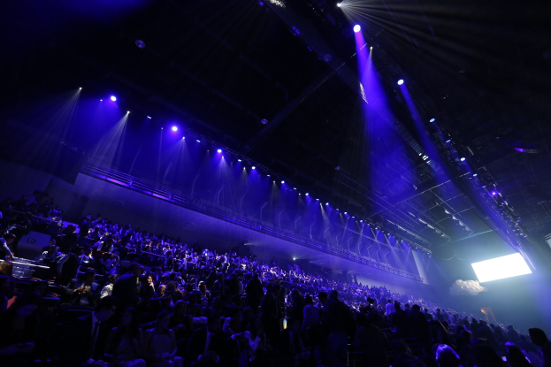 Eine große Menschenmenge vor einer Bühne mit Deckenbeleuchtung und einem Bildschirm auf der rechten Seite.