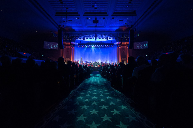Ein großer Zuschauerraum mit Sitzplätzen, eine Bühne mit Musikern und Deckenleuchten und -fernseher, was auf eine Konzerthalle hinweist.