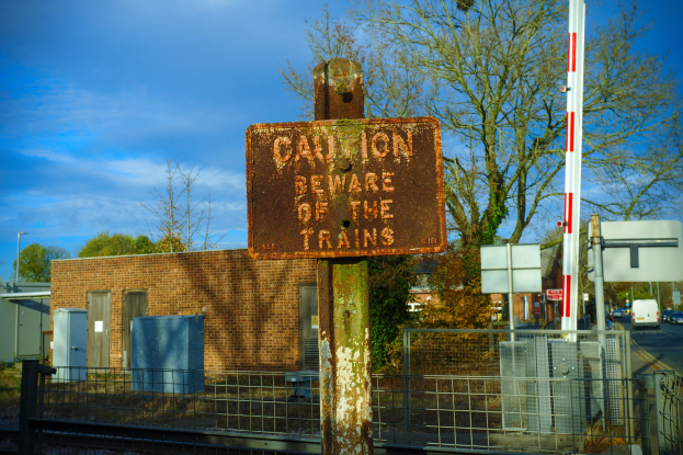 Vorsichtsschild an einem Bahngleiszaun mit Bäumen, Pfählen, einem Gebäude, Containern, Fahrzeugen und einem bewölkten Himmel.