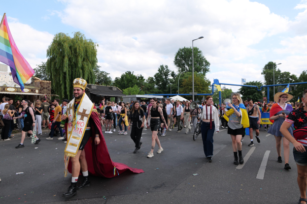 Eine Gruppe von Menschen bei der Pride Parade 2018, einige mit Musikinstrumenten und Mützen, die eine Regenbogenfahne tragen, während sie eine Straße mit Laternen, Bäumen, Schuppen und einem bewölkten Himmel entlanggehen.