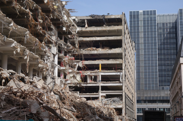 Ein großes Gebäude wird in einer Stadt mit anderen Gebäuden, Fahrzeugen, Straßenelementen, Bäumen und einem klaren Himmel abgerissen.