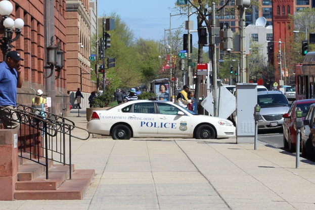 Eine belebte Stadtstraße mit einem geparkten Polizeiwagen, fahrenden Fahrzeugen einschließlich eines Busses und eines Autos, Fußgängern auf dem Gehweg, Gebäuden mit Fenstern und Geländern, Laternen, Bäumen und einem klaren blauen Himmel im Hintergrund.