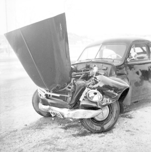 Schwarzes und weißes Foto eines schwer beschädigten Autos am Straßenrand mit eingedellter Motorhaube, zersplitterten Fenstern und offener Fahrertür, die den Motorraum zeigt.
