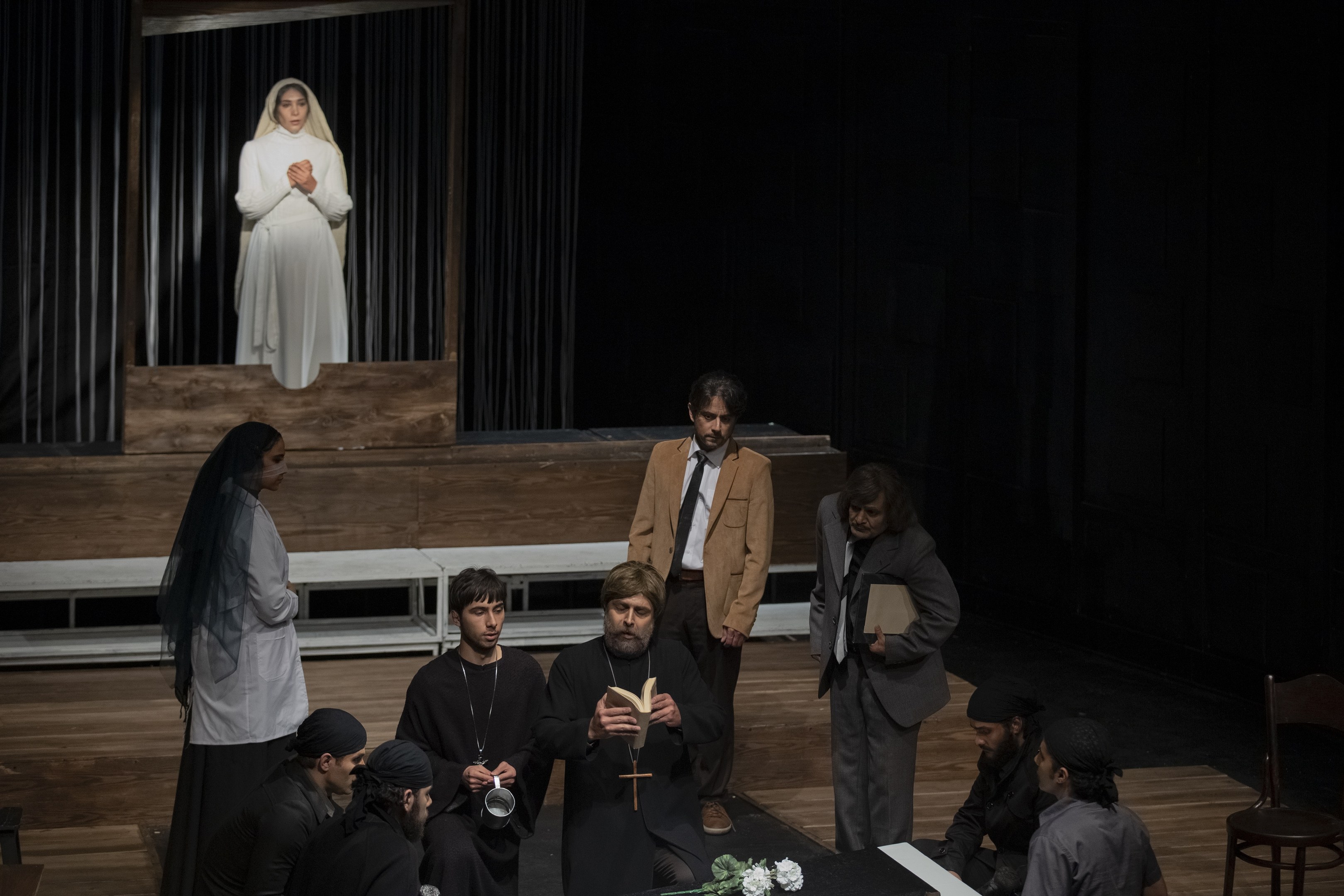 Eine Gruppe von Menschen sitzt um einen Tisch auf einer Bühne, mit einem Mann, der ein Buch in der Mitte hält, umgeben von einer Blumenvase und anderen Gegenständen und Stühlen rechts; Vorhänge und eine Statue einer Frau sind im Hintergrund zu sehen.