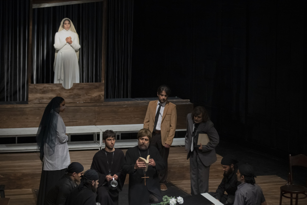 Eine Gruppe von Menschen sitzt um einen Tisch auf einer Bühne, mit einem Mann, der ein Buch in der Mitte hält, umgeben von einer Blumenvase und anderen Gegenständen und Stühlen rechts; Vorhänge und eine Statue einer Frau sind im Hintergrund zu sehen.