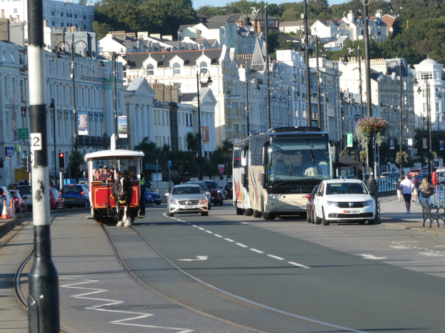 Stadtstraße mit starkem Verkehr, hohen Gebäuden, Fahrzeugen, Fußgängern, Gehwegen, Laternen, Verkehrszeichen, Schildern, Bäumen und einem klaren blauen Himmel.