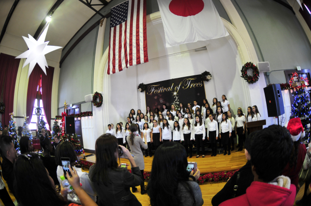 Eine Gruppe von Menschen auf einer Bühne vor einem Publikum mit Weihnachtsdekoration, einem Rednerpult, Lautsprechern, einer Texttafel, Vorhängen, Flaggen und Deckenleuchten.