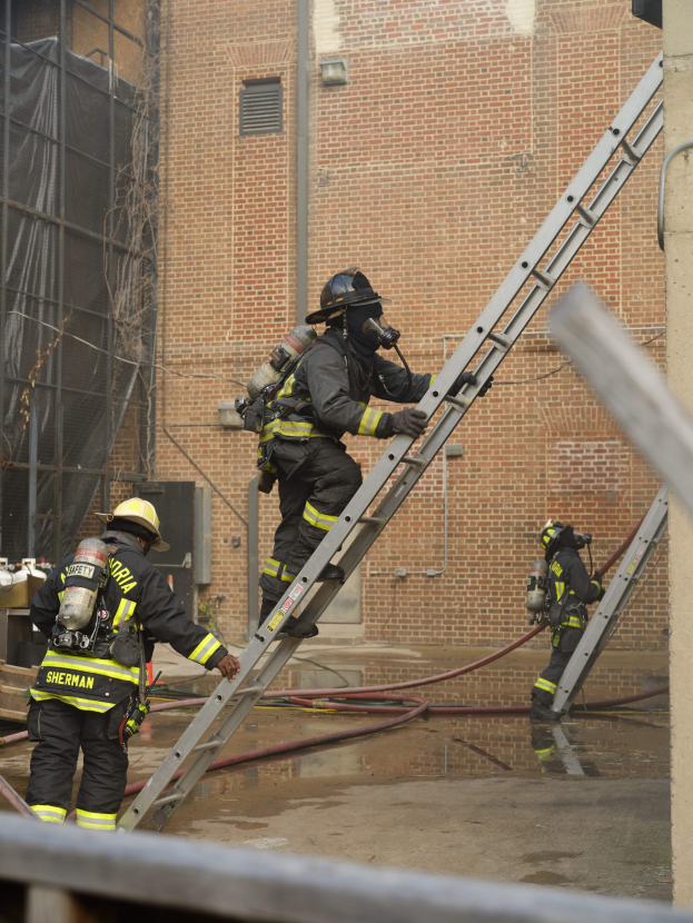 Feuerwehrleute in Helmen und Ausrüstung klettern an einer Leiter vor einem Backsteingebäude mit Rohren auf dem Boden und einer Metallstange unten.