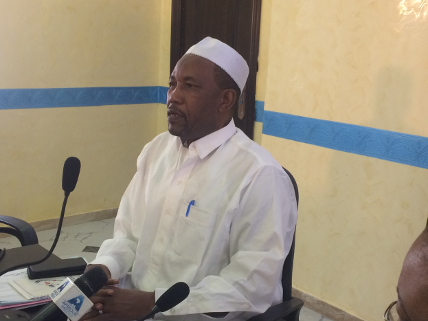 Ein Mann in weißem Anzug und Hut sitzt an einem Tisch mit Mikrofonen, spricht, während Papiere und Gegenstände auf dem Tisch zu sehen sind, mit einem anderen Menschen mit Brille rechts von ihm, in einem Raum mit einer Wand und einer Tür im Hintergrund.