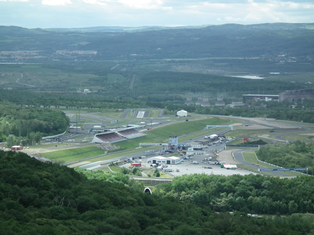 Luftaufnahme einer Rennstrecke mit Fahrzeugen, Gebäuden, Bäumen und Hügeln im Vordergrund und einem bewölkten Himmel im Hintergrund.