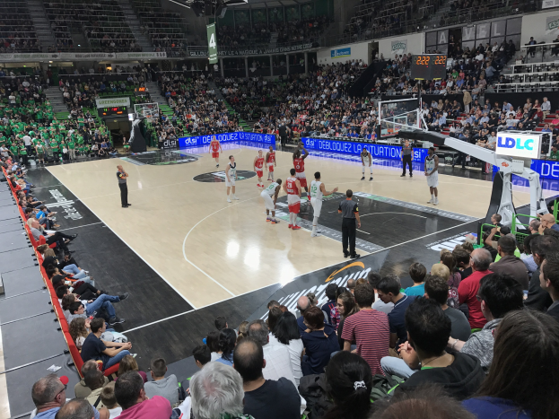 Eine Gruppe von Menschen spielt Basketball in einer großen Arena mit Zuschauern auf Tribünen und Stühlen entlang des Courts.