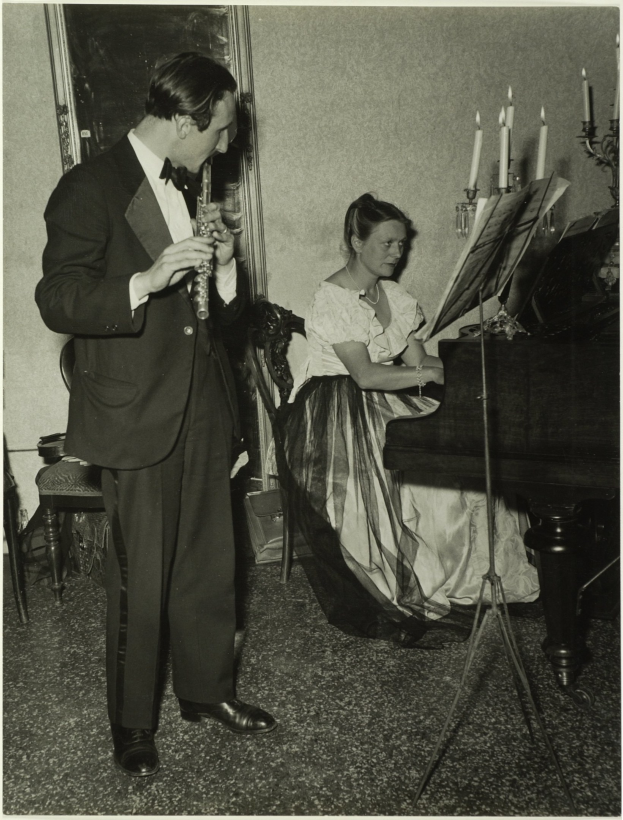 Ein Mann im Smoking spielt Klarinette, während eine Frau am Piano sitzt und Noten auf dem Ständer hat, gerahmtes Kunstwerk an der Wand im Hintergrund sichtbar.