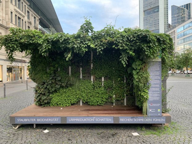 Ein grünes Gebäude mit Efeu an der Seite, umgeben von Bäumen, Fahrzeugen, Pfählen, Schildern und anderen Gebäuden, unter einem bewölkten Himmel, Teil der Staufbilter Biodiversität in Berlin, Deutschland.