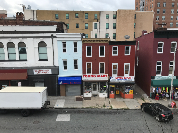 Luftaufnahme einer Stadtstraße in Brooklyn, New York, mit Fahrzeugen, Geschäftsfronten, Werbetafeln, Versorgungsmasten und Drähten vor einem bewölkten Himmel, mit einem zum Verkauf stehenden Gewerbeobjekt.