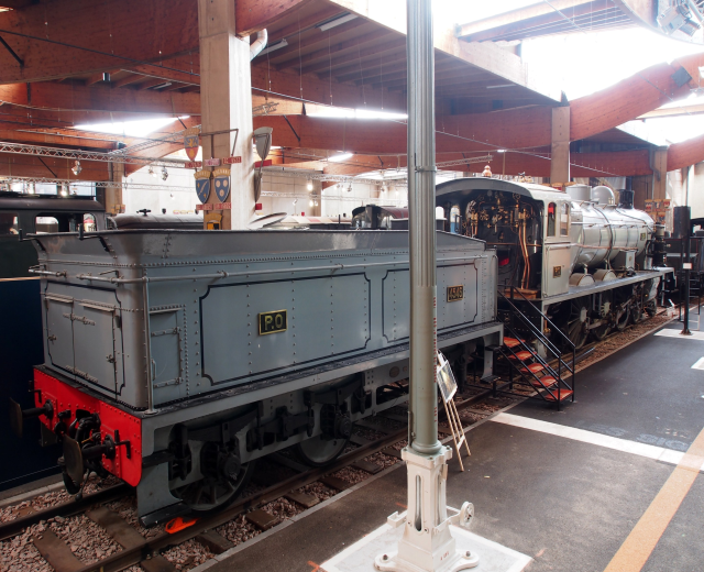 Alte Dampflokomotive in einem Museum ausgestellt, links positioniert mit einer Wand im Hintergrund, umgeben von Schienen, Pfählen und Lichtern.