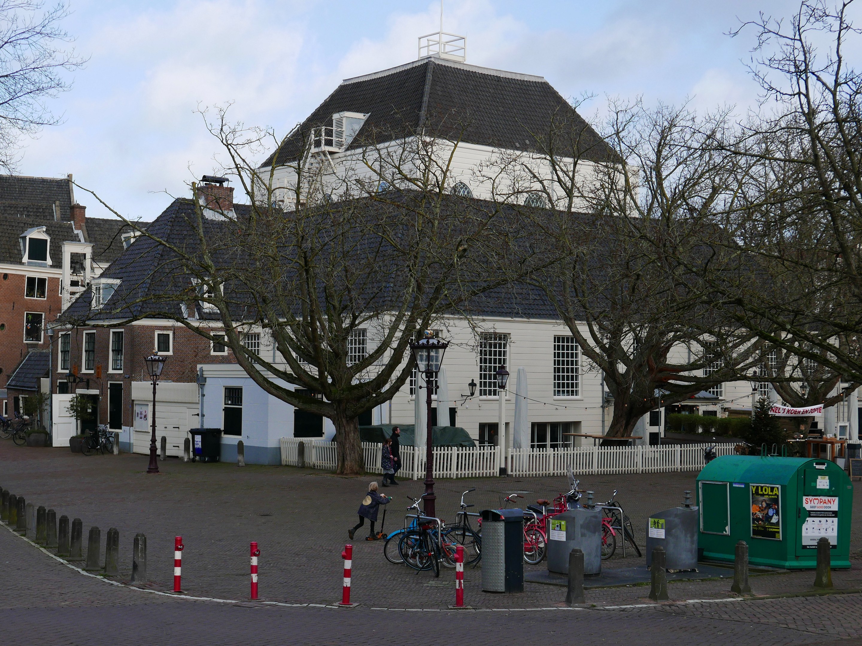 Eine Gruppe von Fahrrädern, die vor einem Rathauses geparkt sind, mit Menschen in der Nähe, umgeben von Bäumen, Pfählen, einem Zaun, Gebäuden und einem bewölkten Himmel.