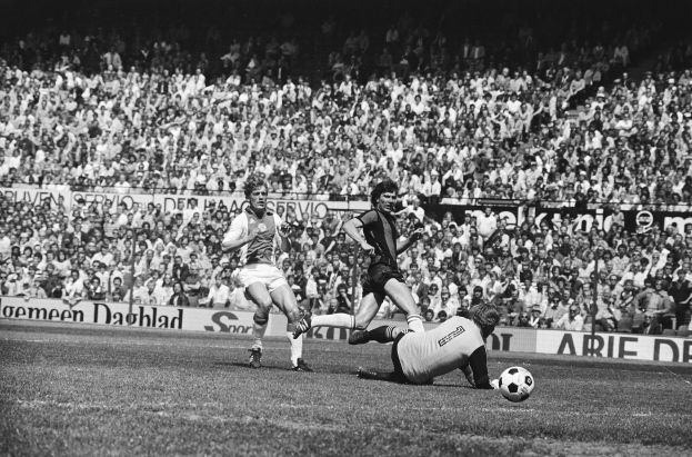 Schwarzes Foto von Männern, die Füßball auf einem Feld mit Zuschauern im Hintergrund und einem Banner "Bundesliga 1978-79 Ajax 1-1 Hannover 96" spielen.