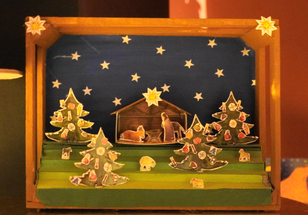 Eine Krippe in einer Holzkiste auf einem Tisch, dekoriert mit Weihnachtsbäumen, Tieren und einem Haus, mit einer Lampe links und einer Wand im Hintergrund.