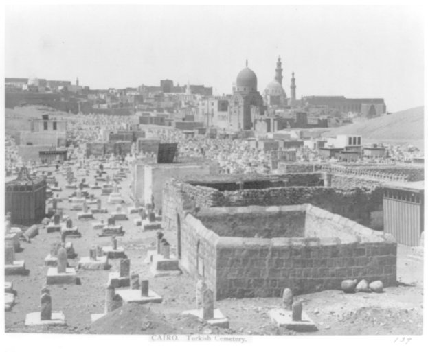 Ein Schwarz-Weiß-Foto eines Friedhofs mit einer Moschee im Hintergrund, umgeben von Gebäuden und einem klaren Himmel, mit Text am unteren Bildrand.