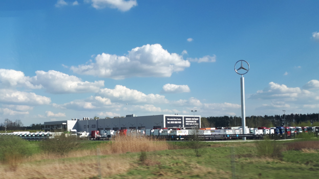 Ein Mercedes-Benz-Werk in Deutschland mit Fahrzeugen auf der Straße, Grünflächen, Gebäuden und einem logogetragenen Pfahl unter einem bewölkten Himmel.
